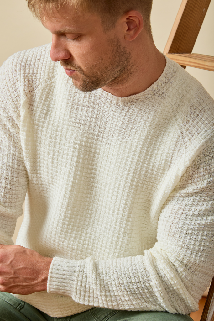 Man wearing a waffle textured cream Merino wool  sweater sitting on a chair.