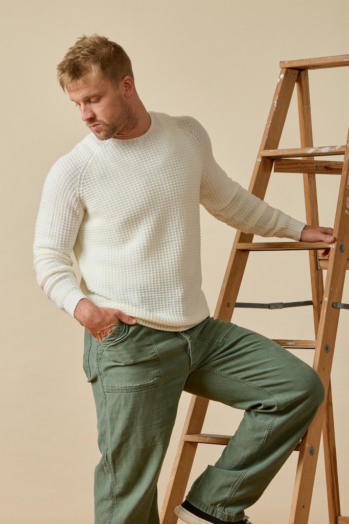 Man in a white Merino wool sweater and green pants standing next to a wooden ladder against a beige background