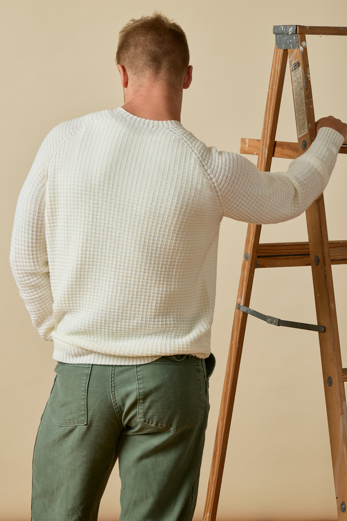Man in a white waffle knit Merino wool  sweater and green pants standing on a wooden ladder against a beige wall.