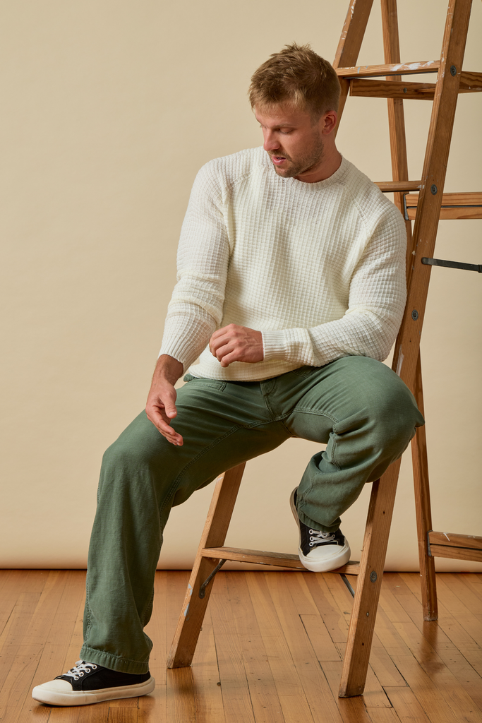 Man sitting on a wooden stool wearing a white Merino wool sweater and green pants.