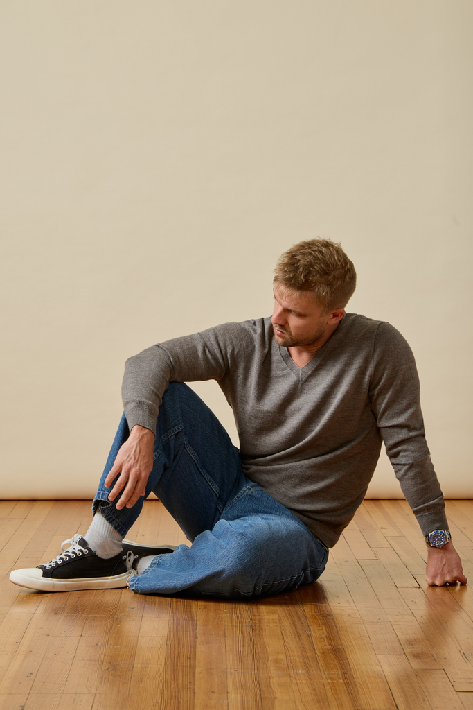Man sitting on a wooden floor wearing a gray Merino wool sweater and blue jeans.