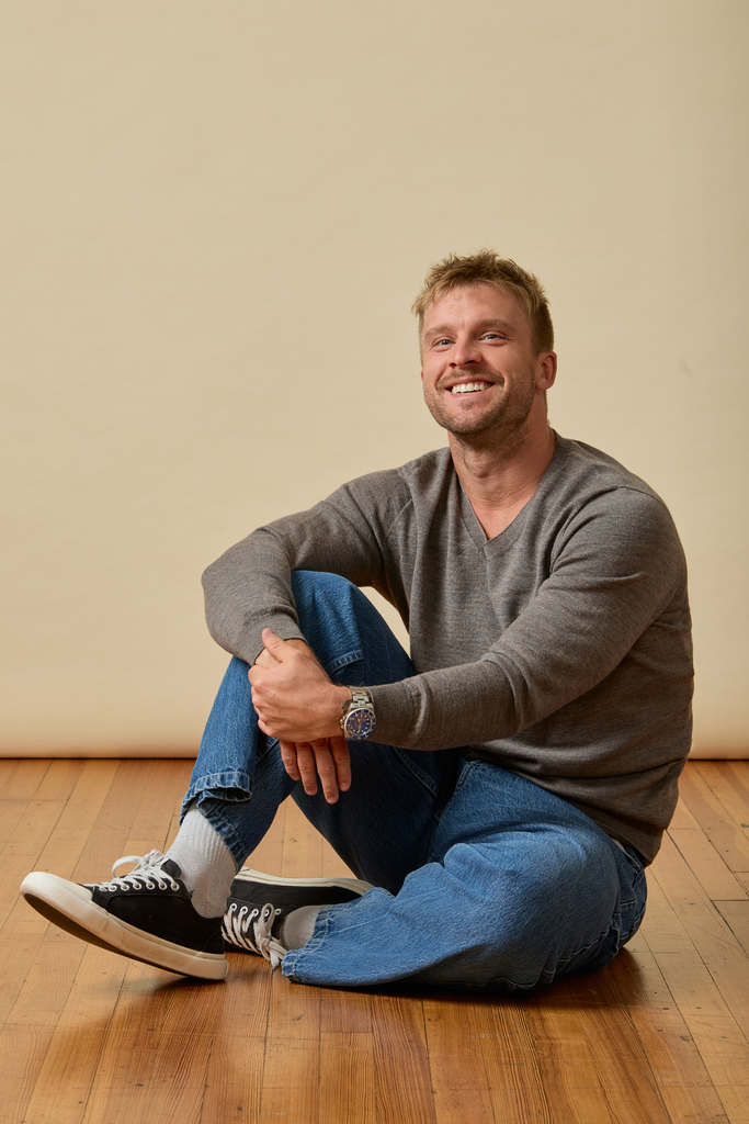 Man sitting on a wooden floor wearing a gray Merino wool sweater and blue jeans.