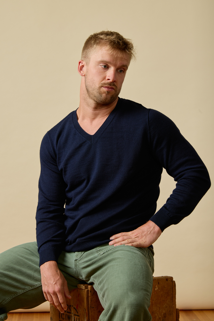 Man wearing a navy Merino wool sweater and green pants sitting on a wooden box against a beige wall.