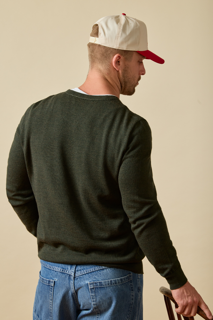 Man wearing a green Merino wool sweater, blue jeans, and a white cap with red brim on a beige background