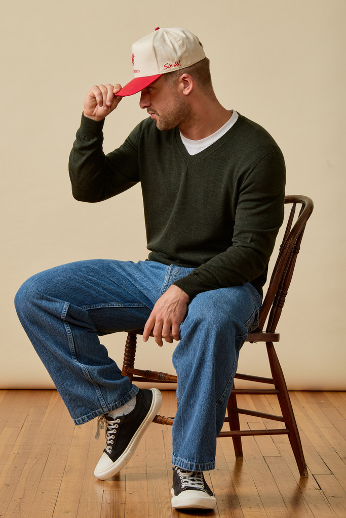 Man sitting on a wooden chair wearing a green Merino wool sweater, blue jeans, and a red and white cap.