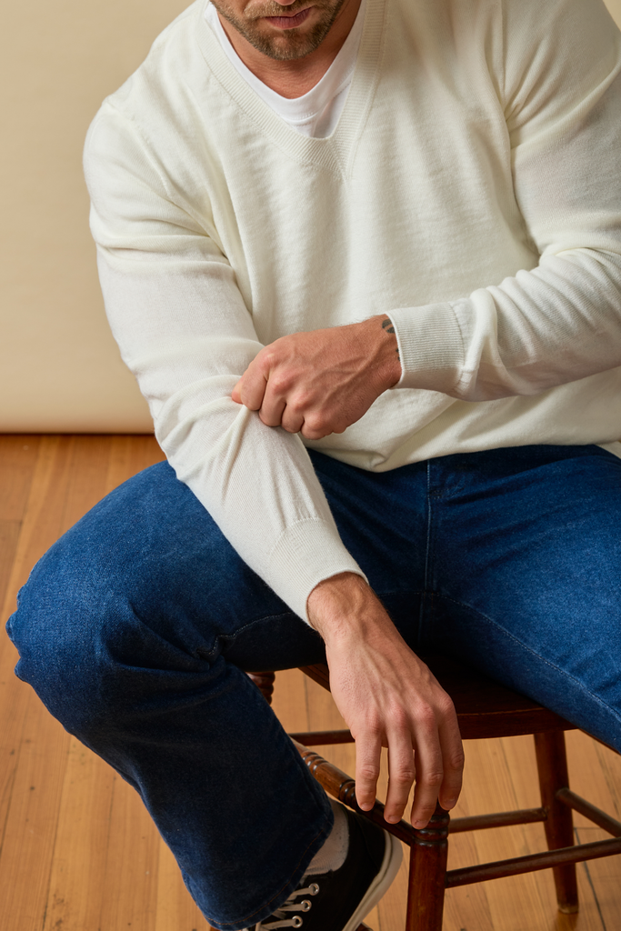 Man wearing a white Merino wool sweater and blue jeans sitting on a wooden stool.
