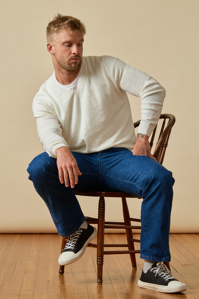 Man sitting on a wooden chair wearing a white Merino wool sweater, blue jeans, and black sneakers.