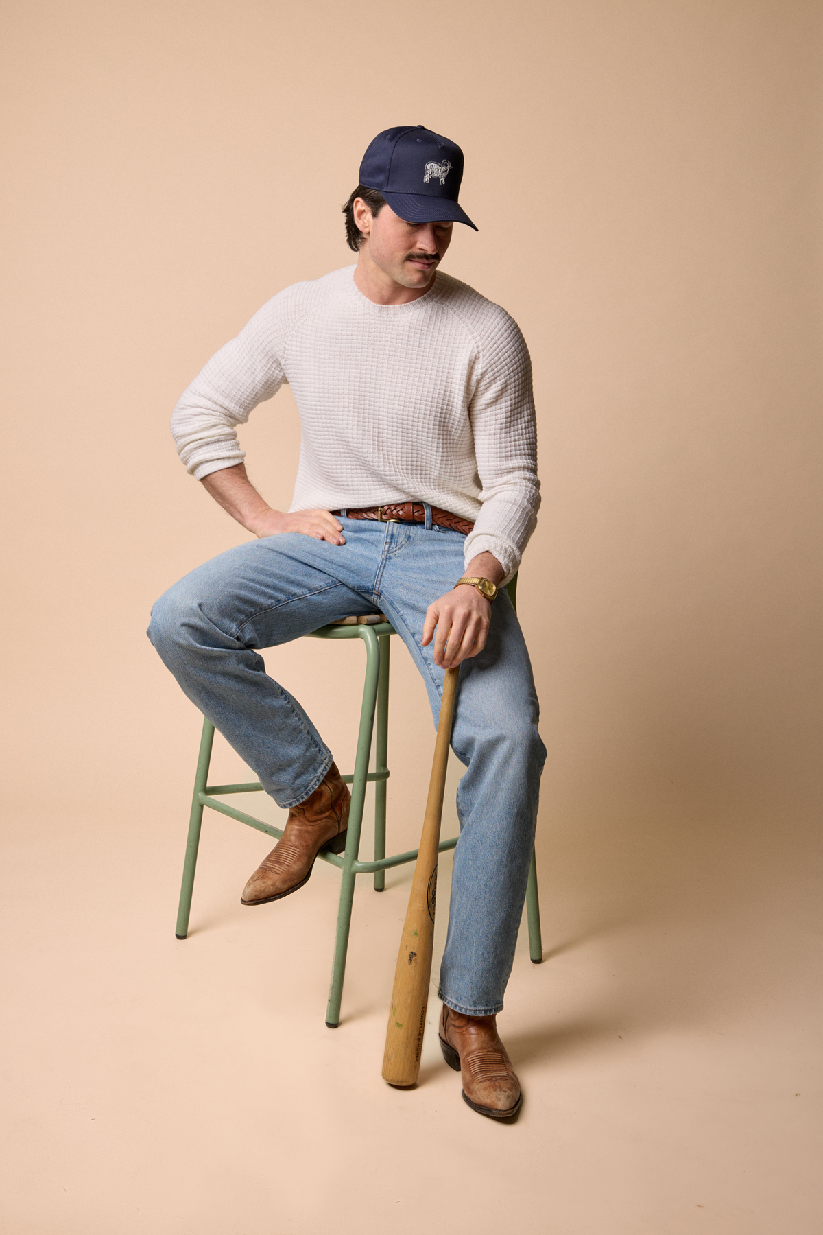 A man wearing a white waffle-knit sweater, light blue jeans, brown leather boots, and a navy baseball cap sits on a green stool holding a wooden baseball bat against a solid beige studio background. #color_naturale