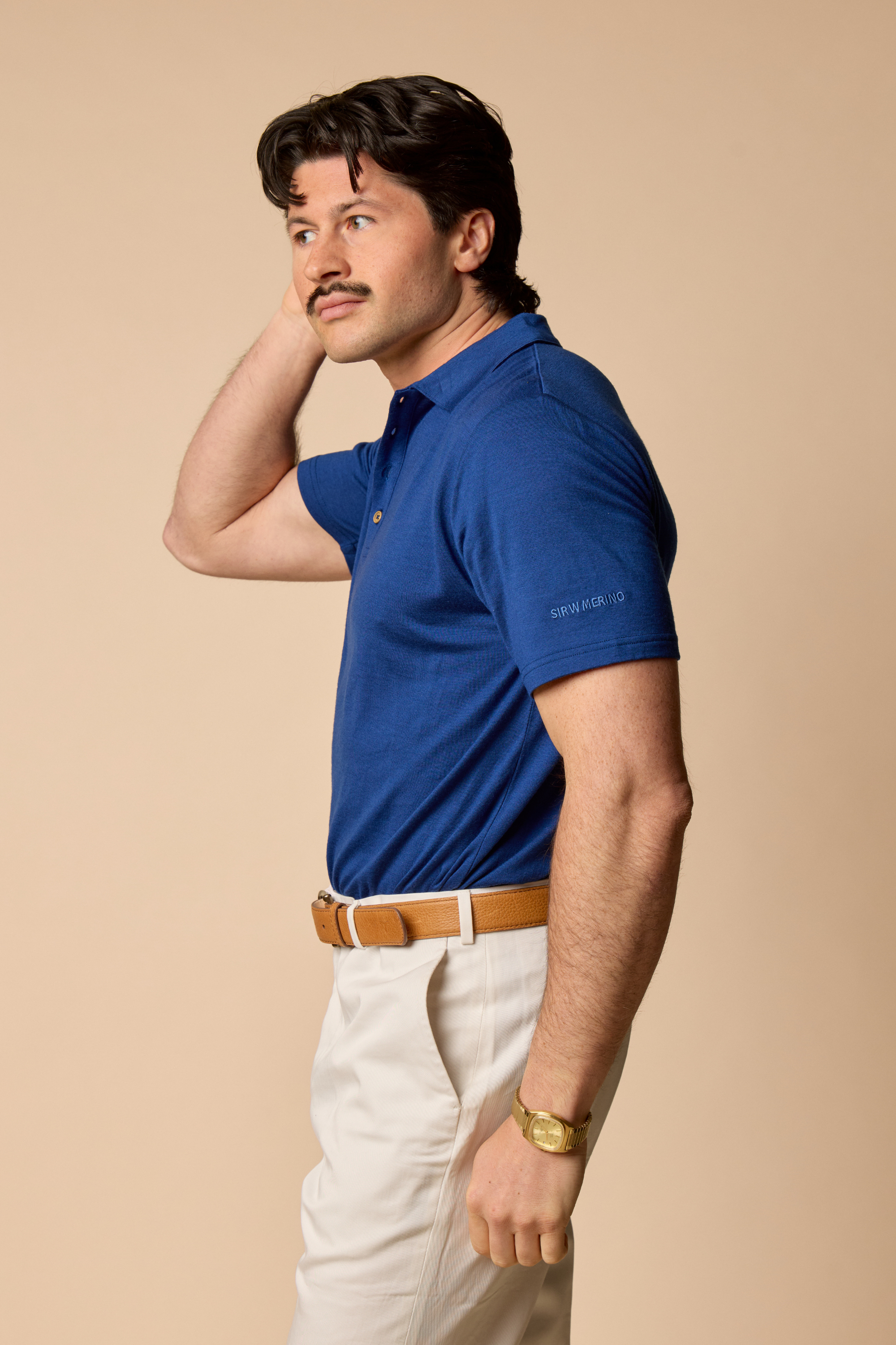 Man wearing a Sir W. Royal blue polo shirt and white pants against a beige background