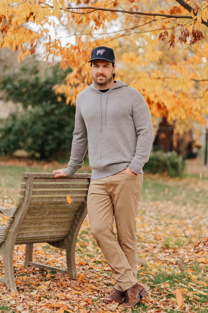 Man in a gray Merino hoodie and beige pants standing next to a wooden bench with autumn leaves and trees in the background.