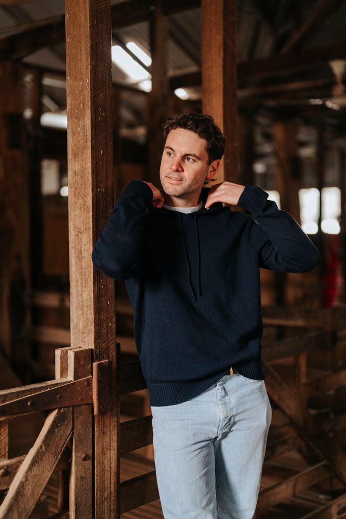 Man wearing a navy blue hoodie and light gray pants standing inside a wooden structure.