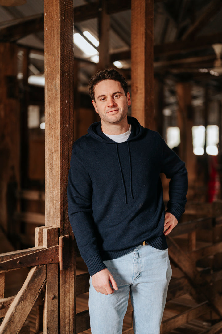 Man wearing a navy hoodie and light jeans standing in a rustic wooden interior.