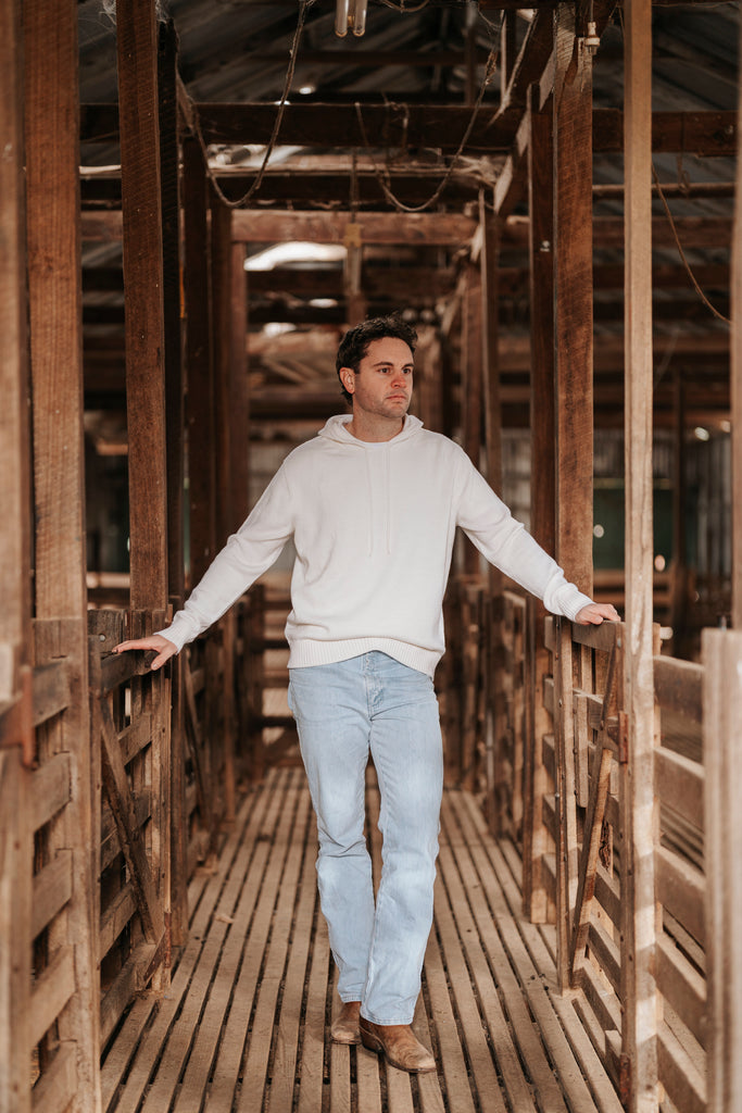 Man wearing a white Merino wool hoodie and light blue jeans standing in a rustic wooden interior.