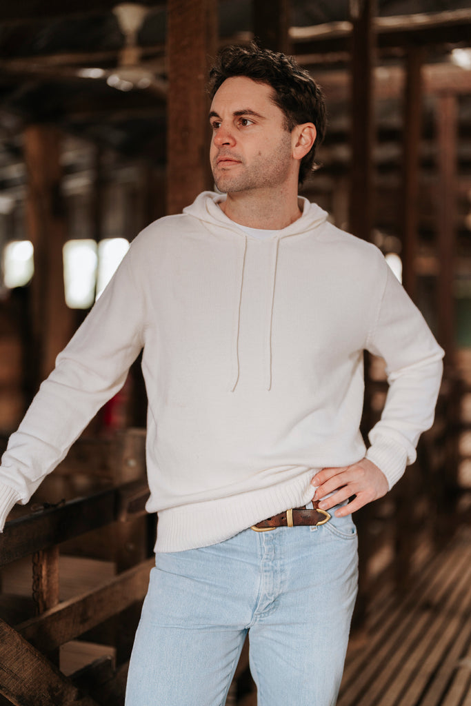Man wearing a white Merino wool hoodie and light blue jeans standing in a rustic wooden interior.