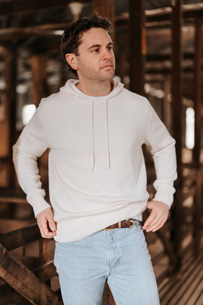 Man wearing a white Merino wool hoodie and light blue jeans standing in a rustic wooden interior.