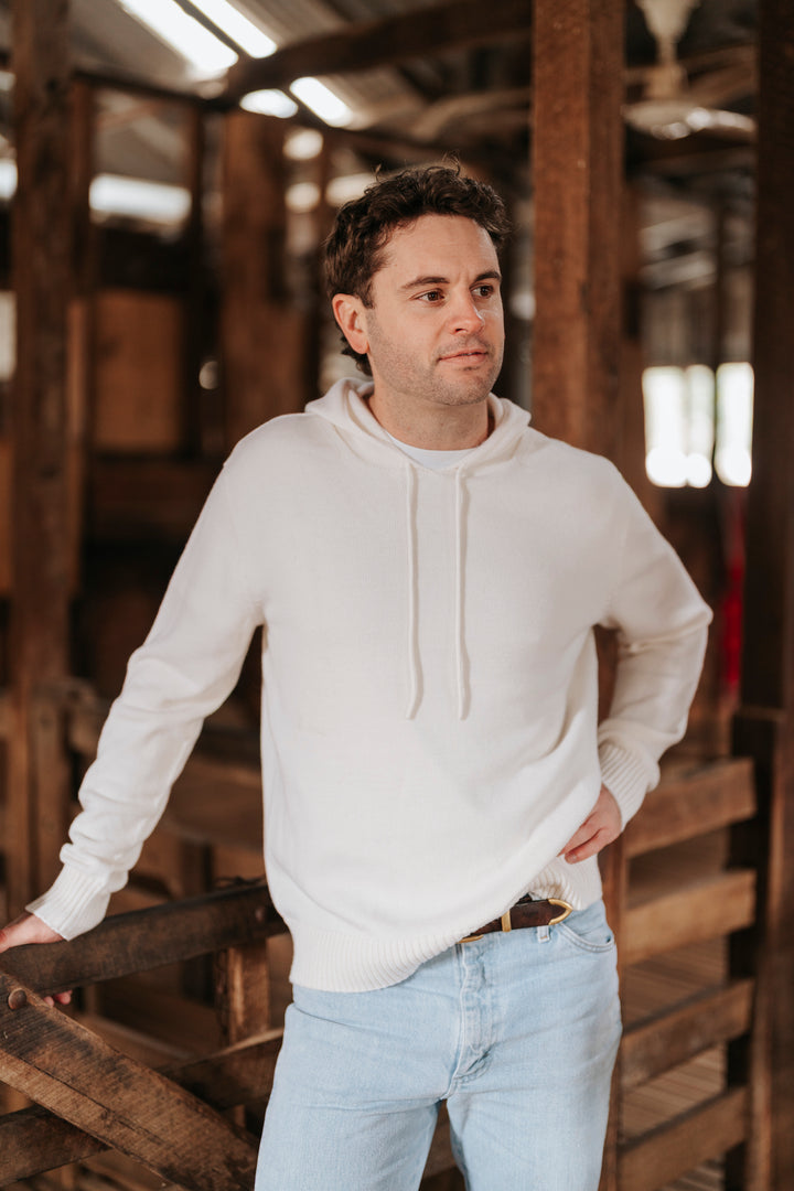 Man wearing a white Merino wool hoodie and light blue jeans standing in a rustic wooden interior.