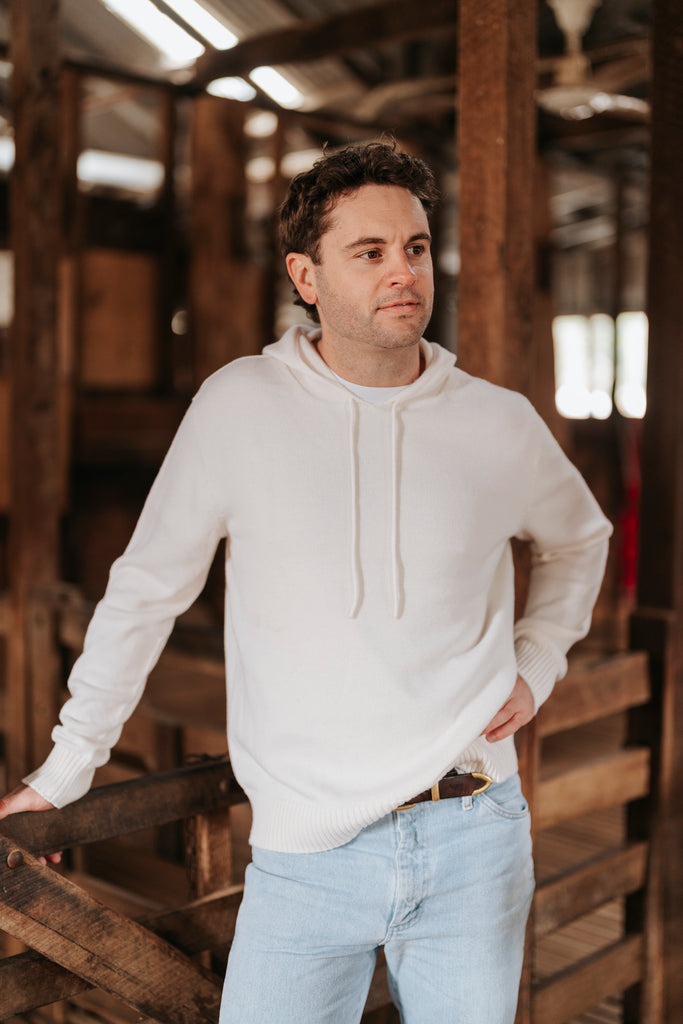 Man wearing a white Merino wool hoodie and light blue jeans standing in a rustic wooden interior.