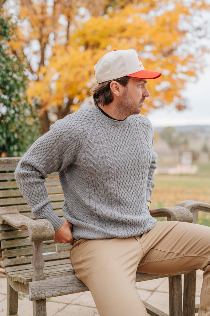 Man sitting on a bench wearing a gray Merino wool sweater and beige pants with a colorful cap in an outdoor setting.