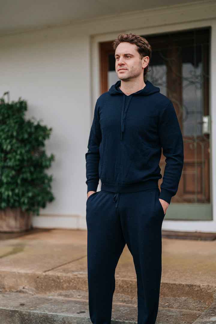 Man wearing a navy blue hoodie and pants standing on a porch.