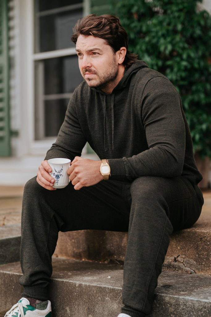 Man sitting on steps holding a mug, wearing a merino hoodie and green sneakers.