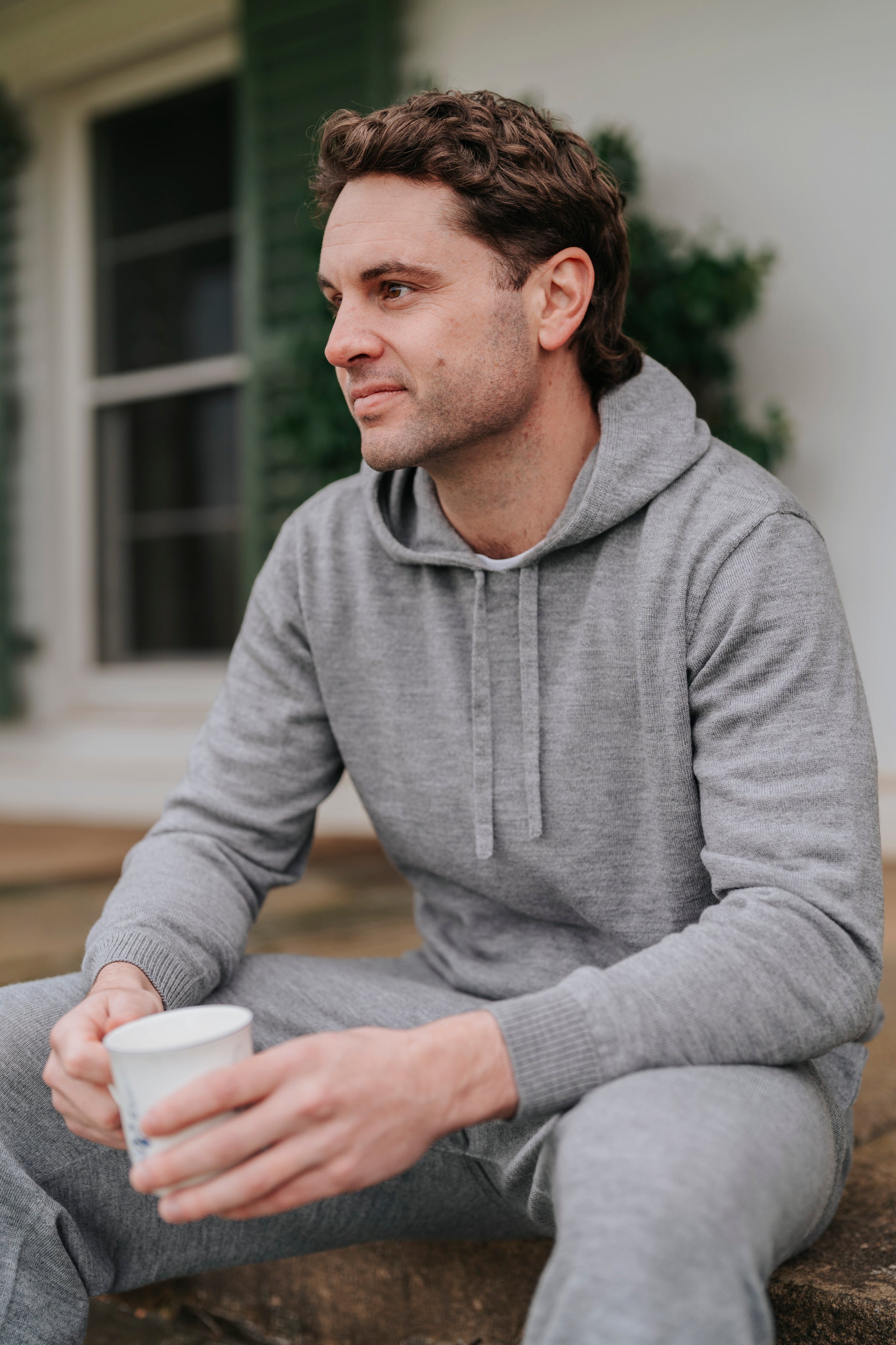 Man sitting outdoors holding a cup, wearing a gray hoodie and pants.