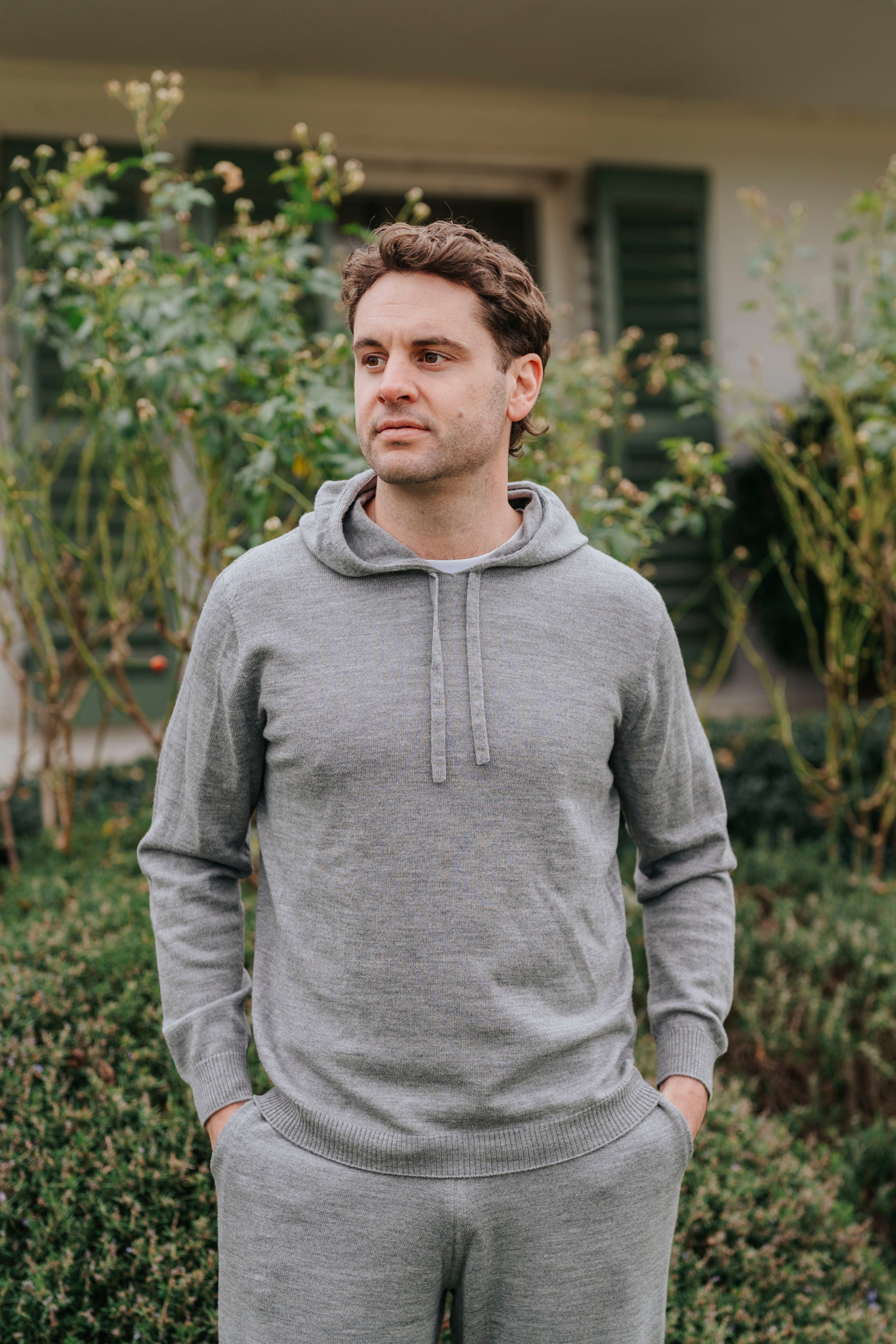 Man wearing a gray hoodie and pants standing outdoors with greenery in the background