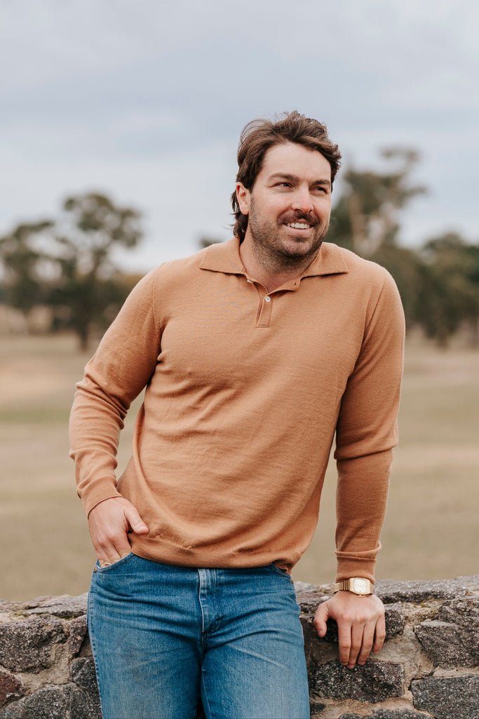 Man wearing a brown long-sleeve shirt and blue jeans standing on a stone wall with a blurred natural background