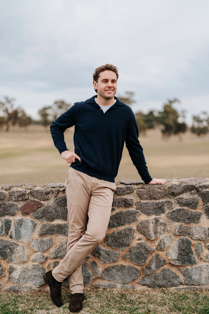 Man leaning against a stone wall outdoors with a field and trees in the background