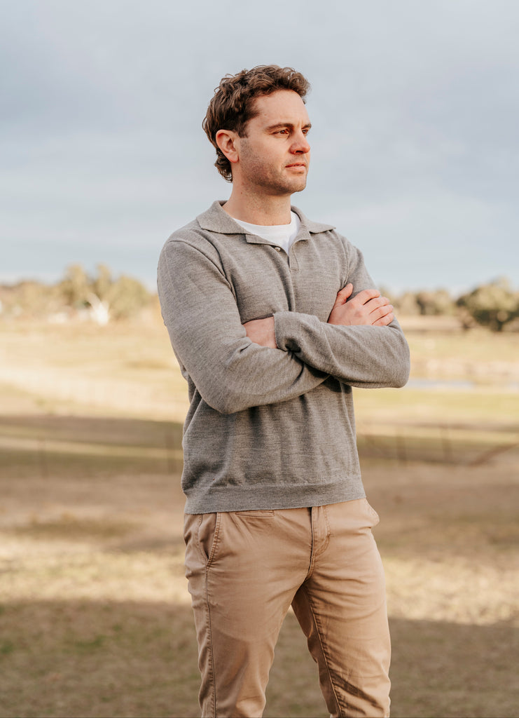 Man standing outdoors with arms crossed, wearing a gray sweater and beige pants.