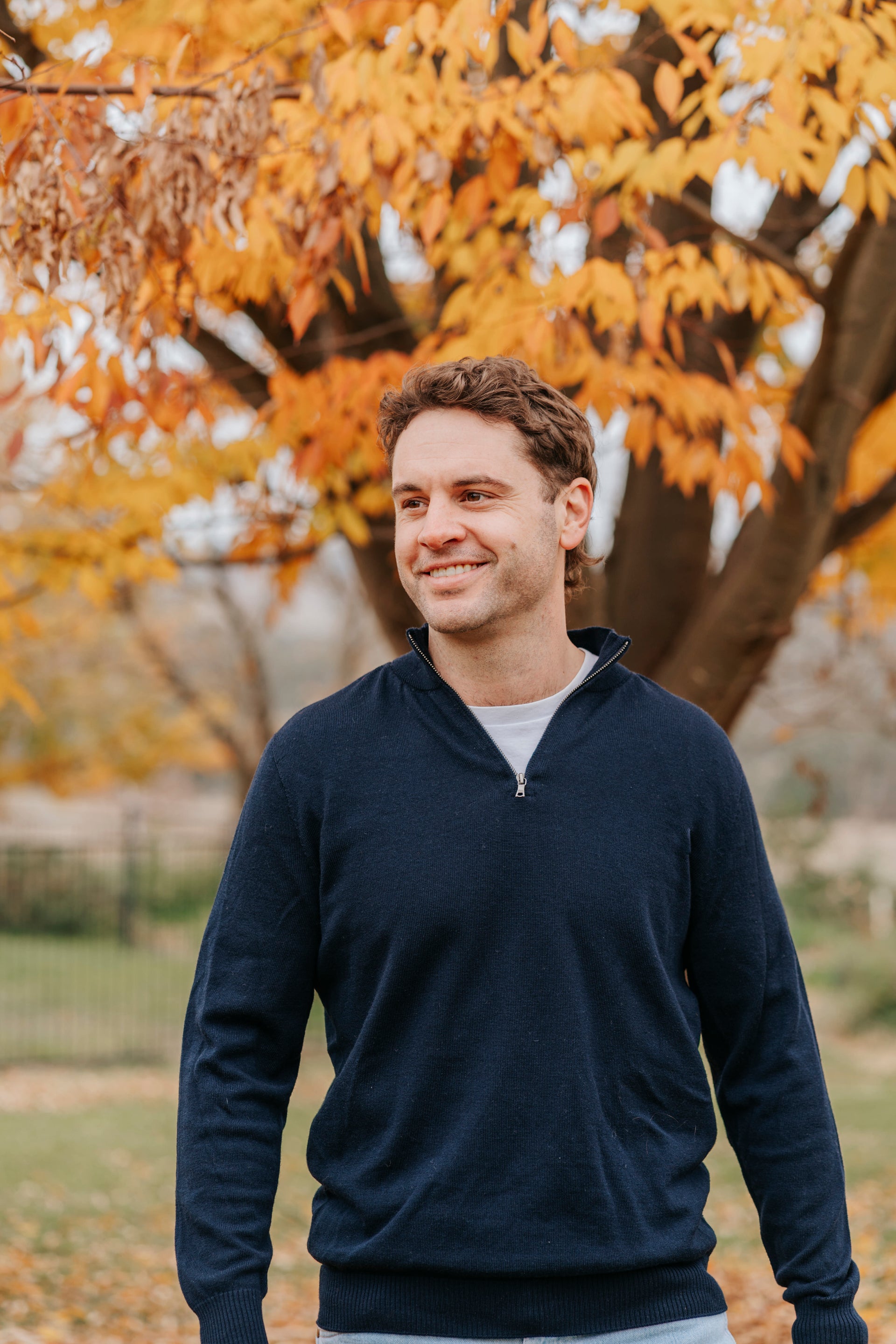 Man wearing a navy blue sweater standing in front of a tree with autumn leaves.
