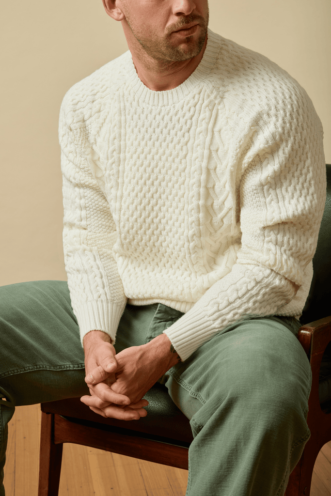 Man wearing a white textured Merino sweater and green pants sitting on a chair.
