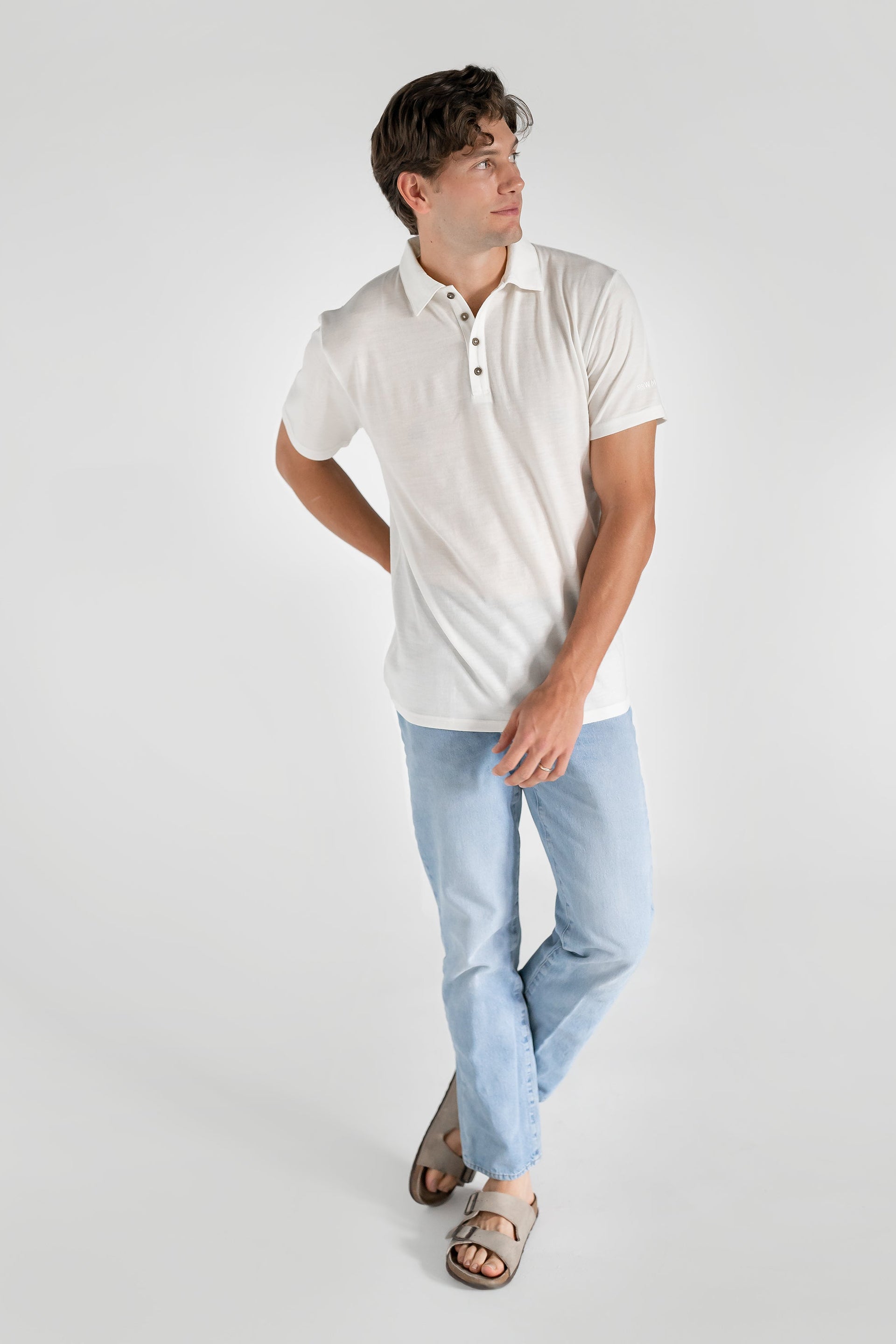 A man wearing a white merino wool polo shirt with a relaxed collar and a four-button placket, paired with light blue jeans.