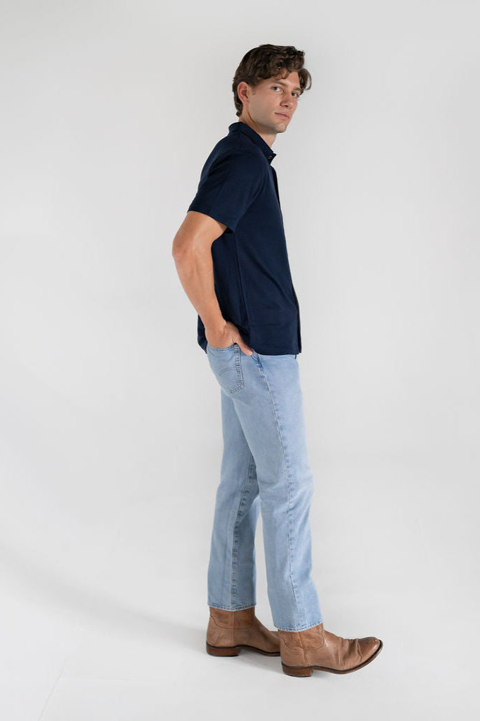 A man wearing a navy blue merino wool polo shirt with a relaxed collar and a 4 button placket, paired with light blue jeans and boots.