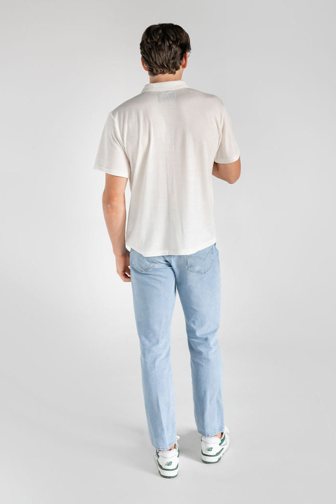 A man wearing a white merino wool polo shirt with a relaxed collar and a four-button placket, paired with light blue jeans.