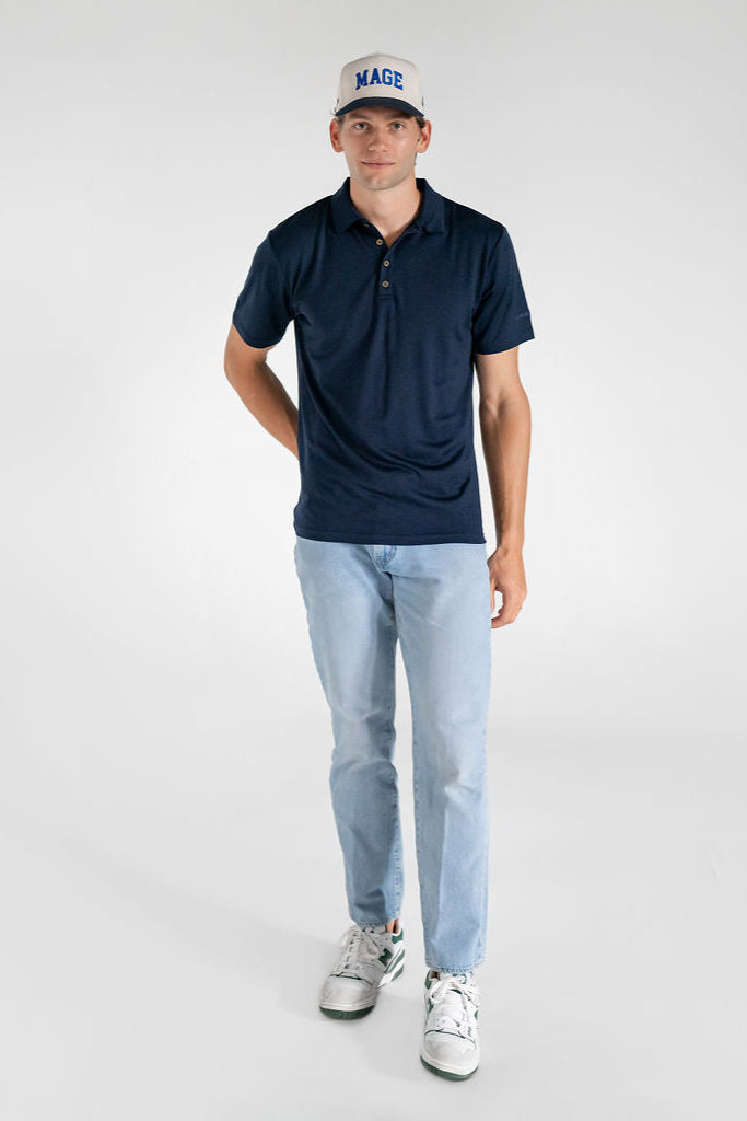 A man wearing a navy blue merino wool polo shirt with a relaxed collar and a 4 button placket, paired with light blue jeans and white sneakers.