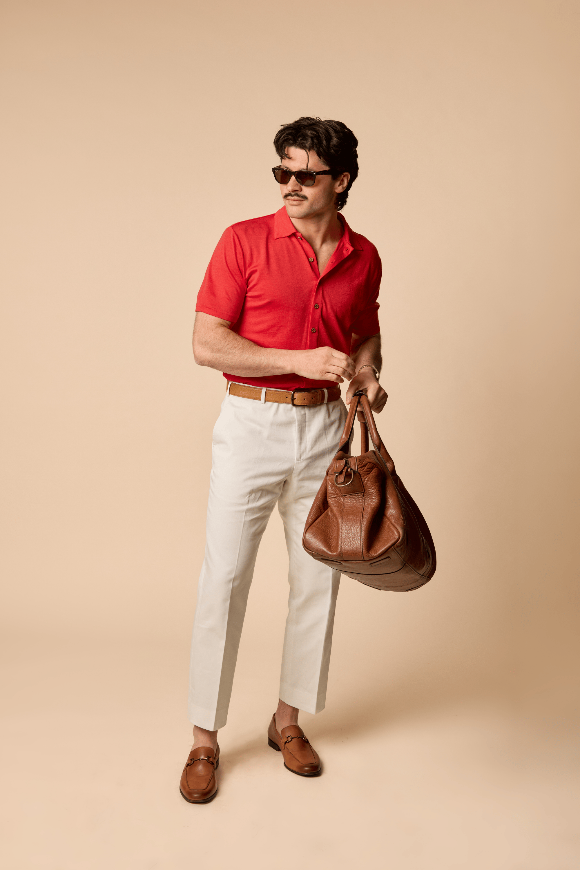 Sir W. Merino Bruce Poppy Red shirt and white pants holding a brown leather bag against a beige background