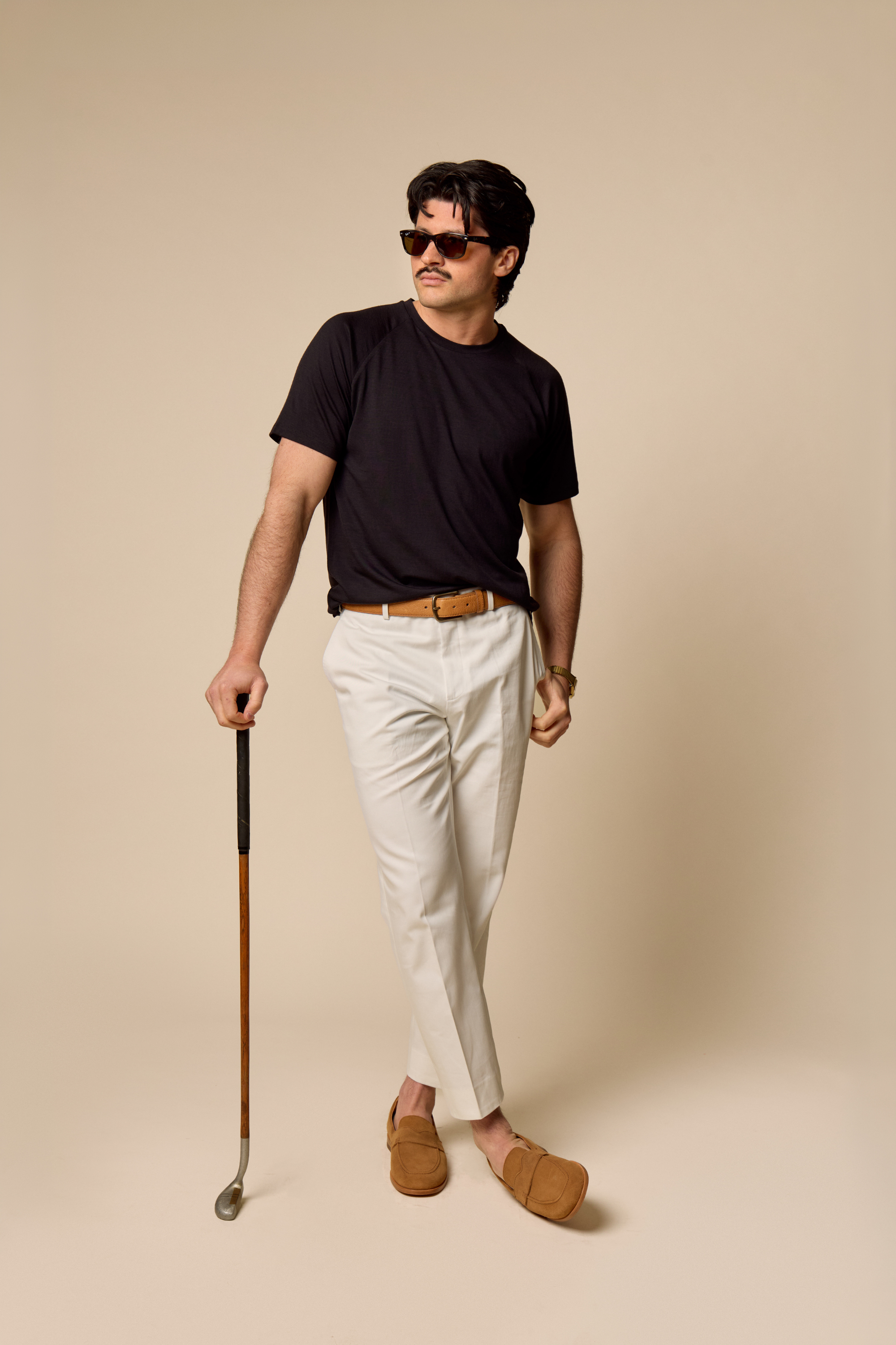 Man in black Sir W. Merino t-shirt and white pants holding a golf club against a beige background