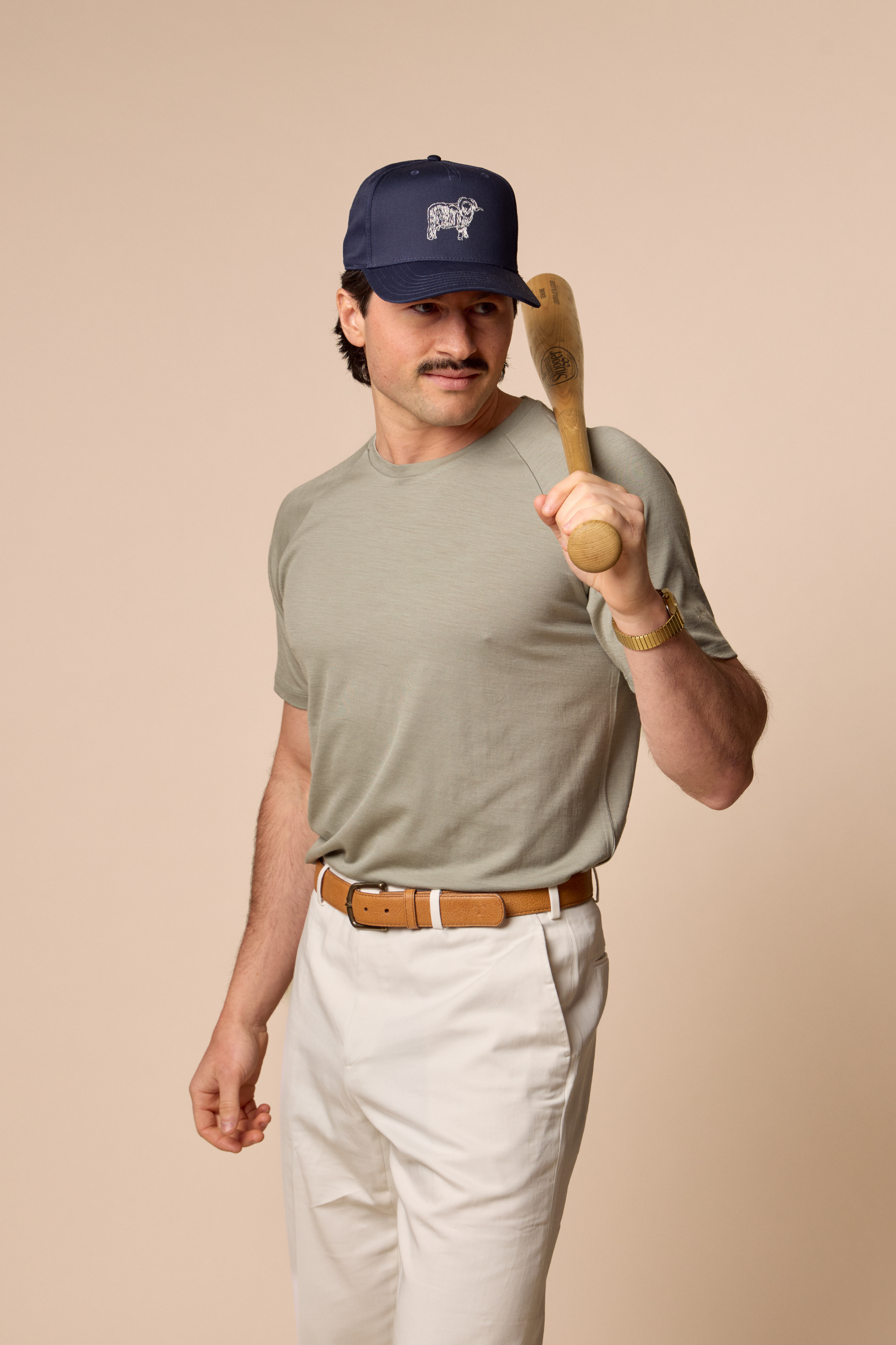 Man wearing Sir W. Cassius athletic tee, holding a wooden baseball bat wearing a cap and casual outfit on a beige background