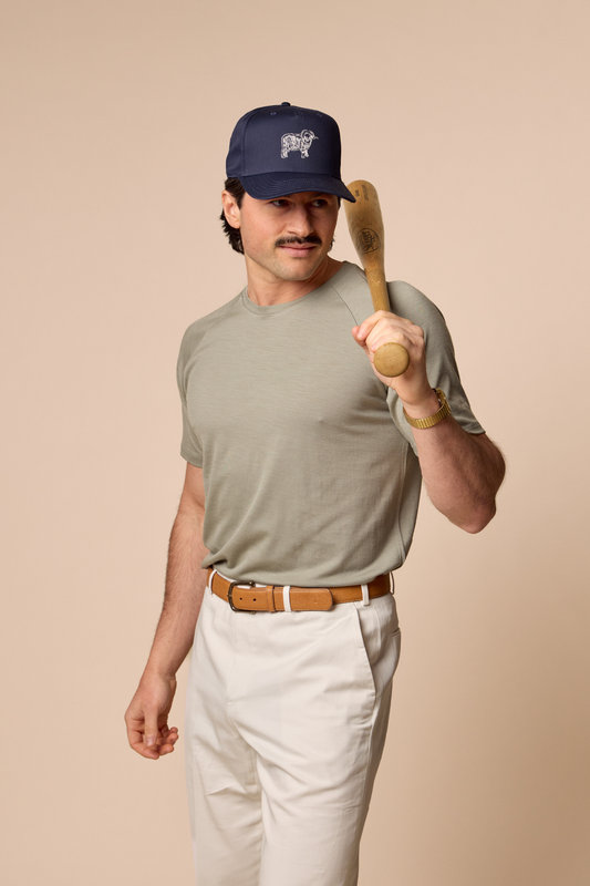 Man wearing Sir W. Cassius athletic tee, holding a wooden baseball bat wearing a cap and casual outfit on a beige background